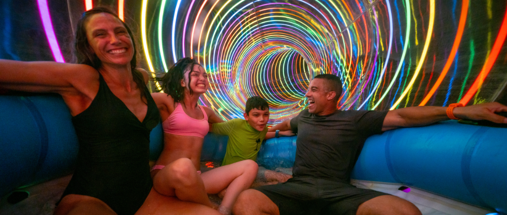 Island H2O Family of four laughing on a water slide raft inside a tunnel with swirling rainbow lights.